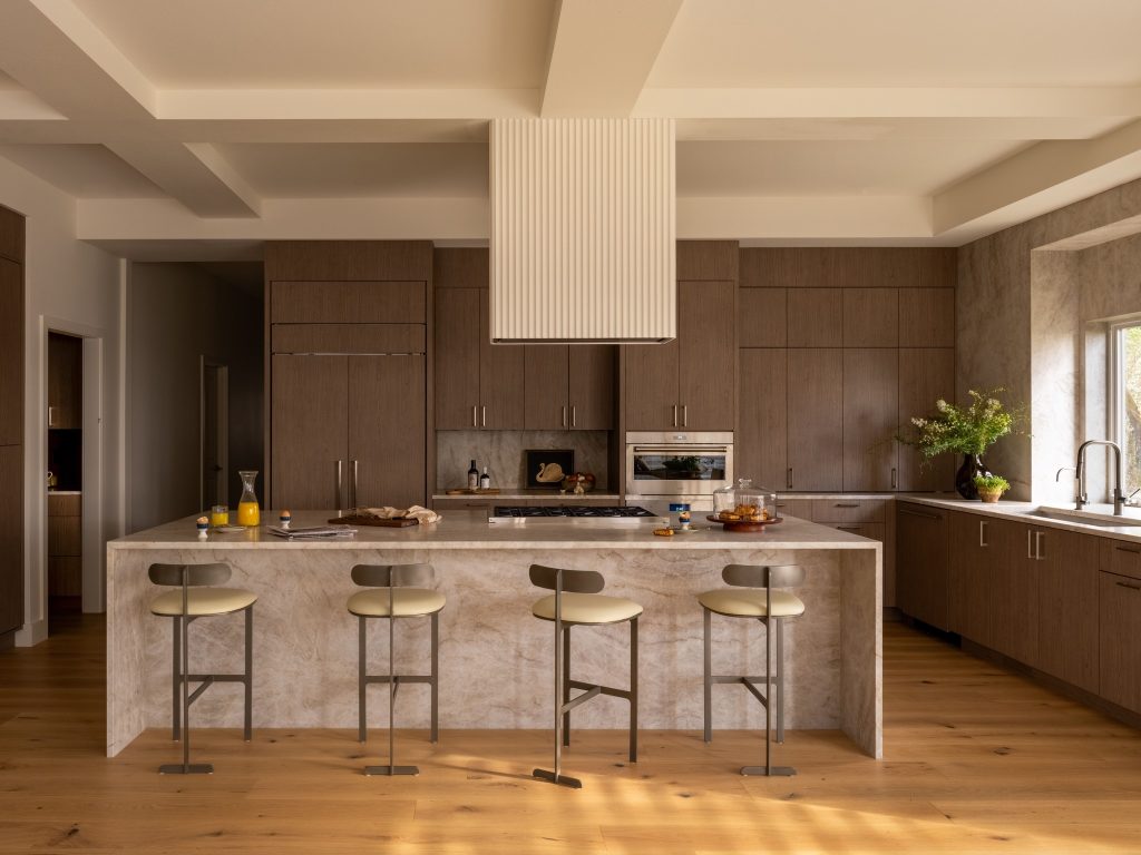 Kitchen with walnut cabinetry, a quartzite waterfall island and four counter stools in warm light.